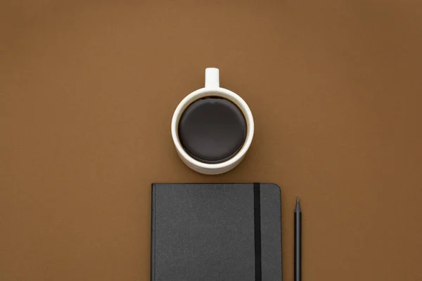 Black notebook, pencil, cup of coffee on the brown background, table top, minimalism working space with copy space.