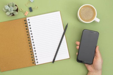 Boş sayfa, kalem, kahve fincanı ve elinde akıllı telefon olan defteri aç. Masa üstü, yeşil arka planda çalışma alanı. Yaratıcı düz yatak.