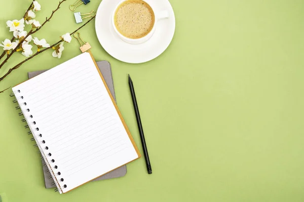 Open notebook with empty page and coffee cup. Spring table top, work space on green background. Creative flat lay.