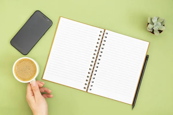Caderno em branco com página vazia, lápis e mão segurando uma xícara de café. Tampo da mesa, espaço de trabalho em fundo verde. Deitado plano criativo . — Fotografia de Stock
