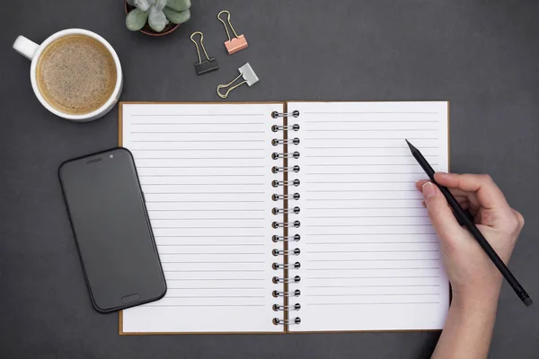 Blank open notebook with empty page, coffee cup and hand holding a pencil. Table top, work space ondark ,textured black background. Creative flat lay. — 스톡 사진