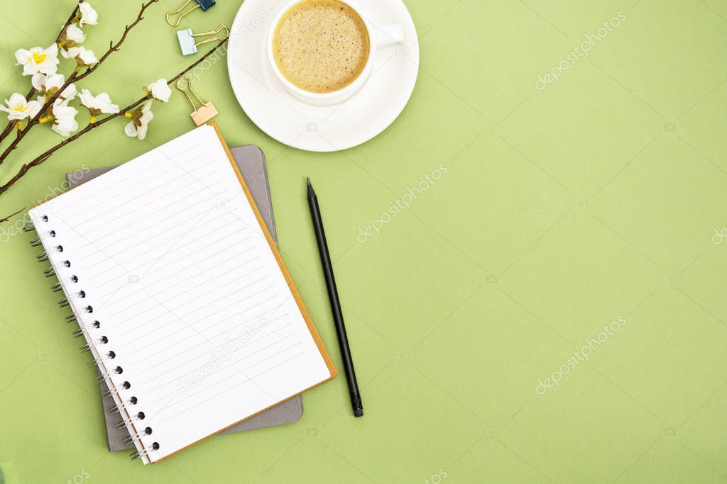 Open notebook with empty page and coffee cup. Spring table top, work space on green background. Creative flat lay.