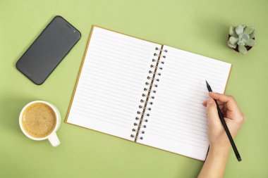 Boş sayfası, kahve fincanı ve elinde kalem olan boş bir defter. Masa üstü, yeşil arka planda çalışma alanı. Yaratıcı düz yatak.