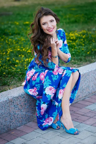 Piękna kobieta uśmiechający się w niebieską sukienkę, siedząc na krawężniku w parku — Zdjęcie stockowe