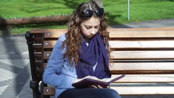Молодая женщина сидит на скамейке в парке и читает книгу — стоковое видео