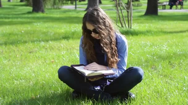 Beautiful young woman sitting in a summer park on the grass and reading a book of the Bible — Stock Video