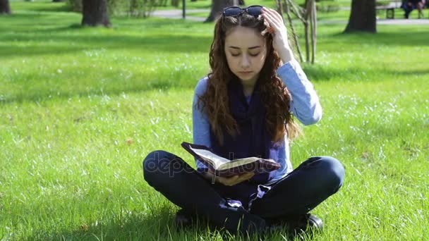 Beautiful young woman sitting in a summer park on the grass and reading a book of the Bible — Stock Video