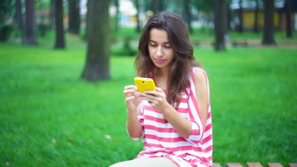 Νεαρή γυναίκα που κάθεται σε ένα παγκάκι στο πάρκο το καλοκαίρι με ένα smartphone — Αρχείο Βίντεο
