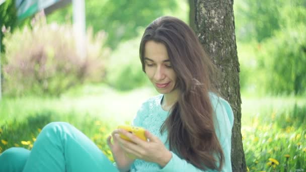 Mulher bonita senta-se sob uma árvore com um smartphone em um parque de verão — Vídeo de Stock