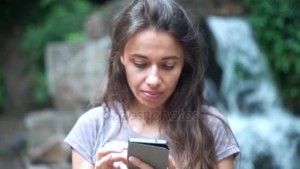 Красивая молодая женщина со смартфоном на фоне водопада — стоковое видео