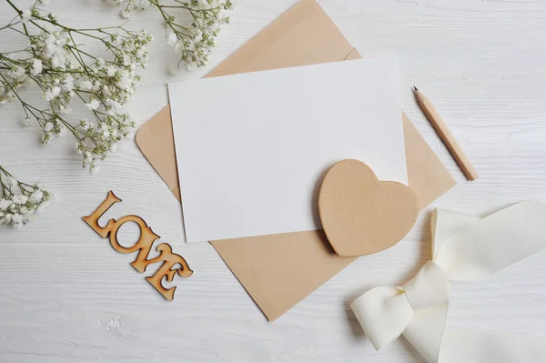 Mock up brief met een liefde vak in de vorm van een hart ligt op een houten witte tafel met gypsophila bloemen, een wenskaart voor Valentijnsdag met een plek voor uw tekst. Foto plat lag, bovenaan weergeven — Stockfoto