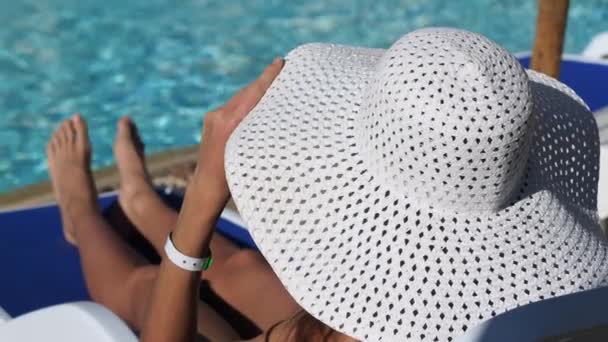 黒い水着を着た幸せな若い女性と白い帽子はホテルのラウンジャーに横たわっています。プールのそばで休もう。海の上のリゾート。Prores 3840x2160の4kビデオ — ストック動画