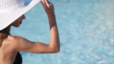 Yakın plan. Bulanık su arka planı. Büyük beyaz şapkalı genç bir kadın havuzun yanında oturuyor ve şapkasını saklıyor. Mesajlaşmak için yer var. 4k video Prores 3840x2160