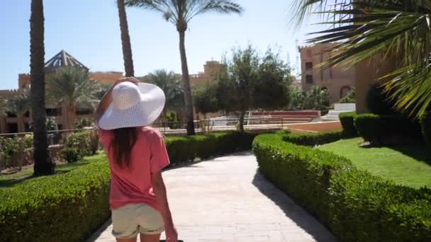 Hermosa mujer en gran sombrero blanco y bolsa de viaje rosa caminando por el territorio del hotel. Camina por el callejón con palmeras. Vídeo HD en ProRes 1920x1080 — Vídeos de Stock