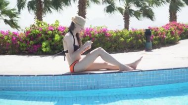 Plajdaki siyah mayo giymiş kadın güneş kremi ya da güneş kremi sürmüş. Yüzme havuzunun yanında oturuyor ve bacağına krem sürüyor.