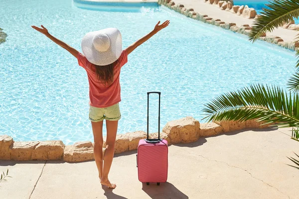 Viagens, férias de verão e conceito de férias - Mulher bonita levanta as mãos perto da área da piscina do hotel com mala rosa — Fotografia de Stock