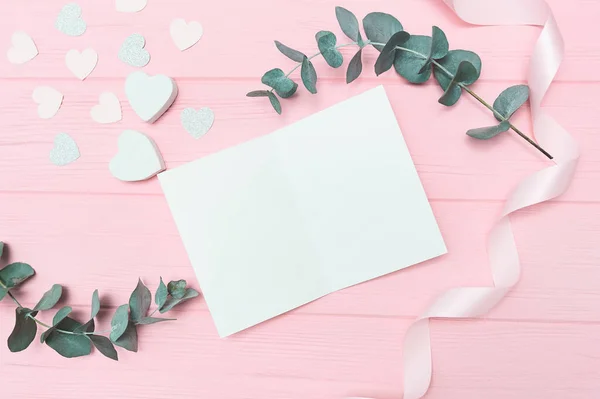 Día de San Valentín o boda escena de maqueta con tarjeta en blanco, hojas de eucalipto marco corazones de papel confeti y fondo rosa de madera, espacio vacío para su texto, vista superior plano laico — Foto de Stock