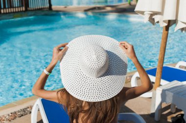 Tropik bir tatil köyünde güneşlenen güzel bir kadın, yaz tatilinin tadını çıkarıyor. Elleri havuz kenarında güneşlenen şapkalı bir kız.