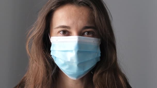 Close-up of a young woman in a protective medical mask, she looks directly at the camera. Concept of health and safety life, N1H1 coronavirus, virus protection, pandemic — Stock Video