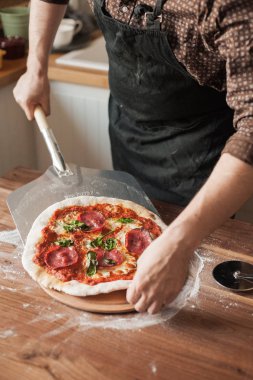Önlüklü bir adam tahta bir masaya domates salçası, salam, peynir ve fesleğenli hazır pizza koyar.