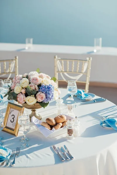 Wedding decor of tables on the island of Santorini in gold, blue and white colors with a bouquet of roses and a gold frame