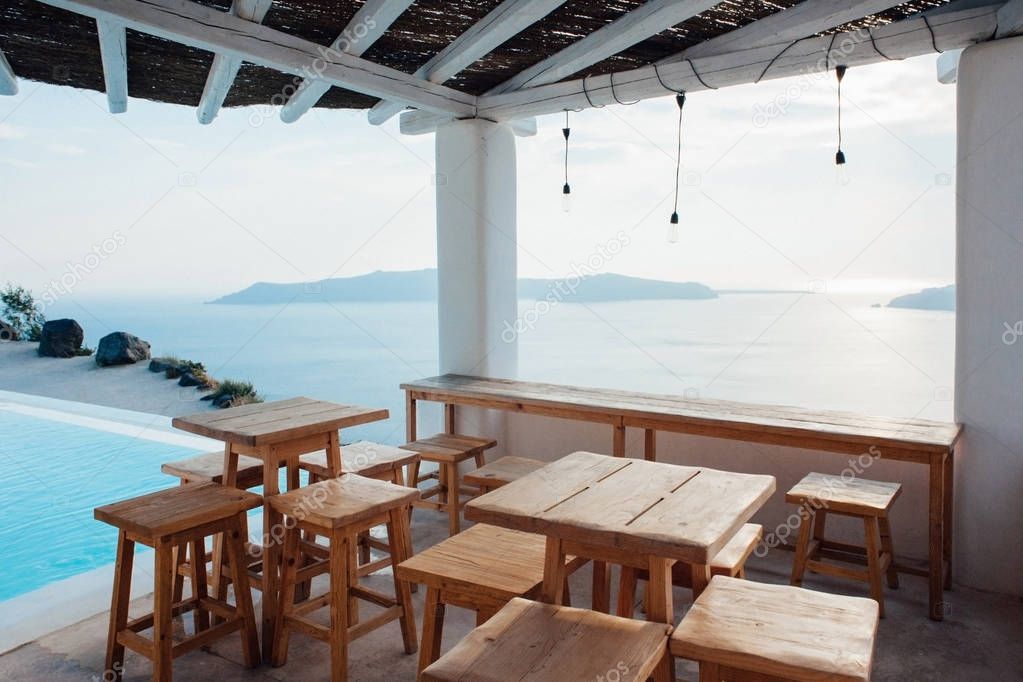 Terrace under the roof with wooden tables and chairs overlooking the sea next to the swimming pool in Santorini island