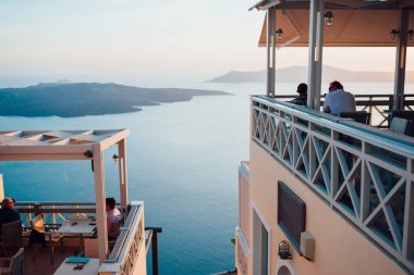 Santorini Adası Deniz, gökyüzü ve geri bir kafe ile büyük bir boş alan, planda yatay oturan insanlar ile adalar üzerinde pembe-mavi günbatımı görünümü