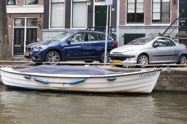 Amsterdam 'da bir kanal boyunca beyaz bir tekne yanaşmış, yakınlarda iki tane park edilmiş araba var. Geleneksel tekne ve modern araçların bu birlikteliği şehirde eşsiz bir ulaşım karışımı sergiliyor.