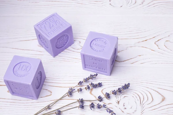 Savon de lavande naturel de couleur lilas sous forme cubique avec inscription en français : Savon de lavande de Marseille, 100 grammes, fleurs de lavande sur une table en bois blanc, style rustique — Photo