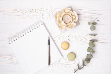 Boş bir albüm, metal bir kalem, okaliptüs filizi, badem kurabiyesi ve beyaz ahşap bir masada çiçek şeklinde bir şamdan.