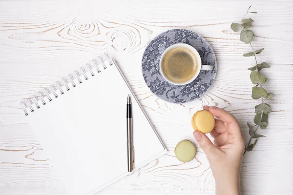 Женская рука держит макарон, чашку кофе и чистый дневник с весной и ветку эвкалипта на белом потрепанном деревянном столе. Модная квартира — стоковое фото