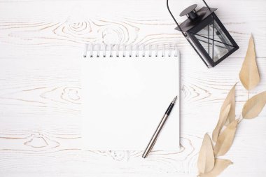 Boş bir albüm ve metal kalem, bej yapraklar, beyaz ahşap bir masada eski bir fener şeklinde siyah bir şamdan.