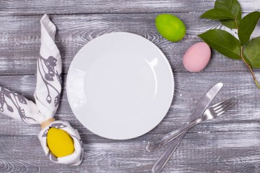Paskalya kompozisyonu boş bir beyaz tabak ve peçeteye sarılı tavşan kulağı, çatal bıçak takımı, renkli Paskalya yumurtaları ahşap gri bir masada bahar yaprakları. Düz yatıyordu
