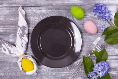 Paskalya kompozisyonu boş bir siyah tabak ve peçeteye sarılı tavşan kulağı, çatal bıçak takımı, renkli Paskalya yumurtaları ahşap gri bir masada bahar çiçekleri. Düz yatıyordu.
