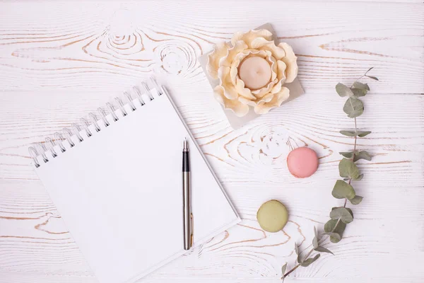 Top view of an empty album and a metal pen with a feather, sprigs of eucalyptus, macaroons and a candlestick in the shape of a flower on a white wooden table, place for text.
