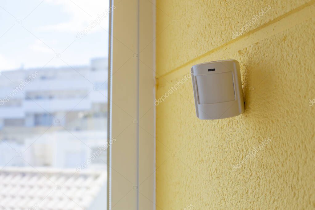 Motion sensor or detector for security system on a yellow wall by the window, indoors. Place for text.