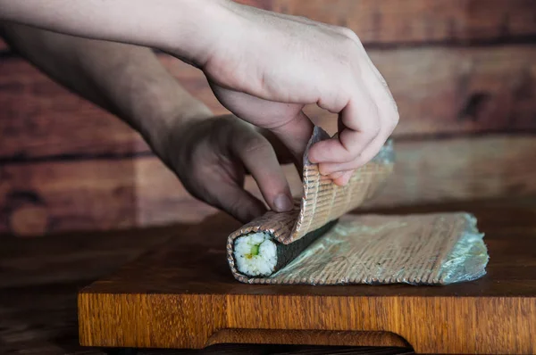 Process of making maki sushi. Cook chef hands preparing rolls with cheese, cucumber and sesame seeds on wooden board with romaji maki sudare