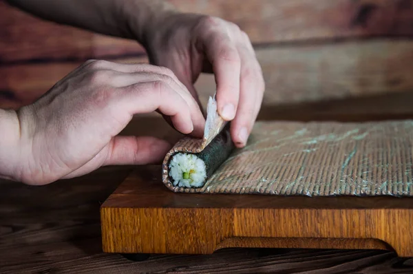 Process of making maki sushi. Cook chef hands preparing rolls with cheese, cucumber and sesame seeds on wooden board with romaji maki sudare