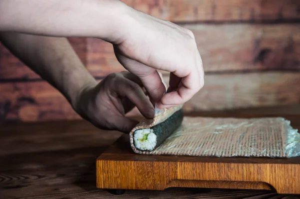 Process of making maki sushi. Cook chef hands preparing rolls with cheese, cucumber and sesame seeds on wooden board with romaji maki sudare