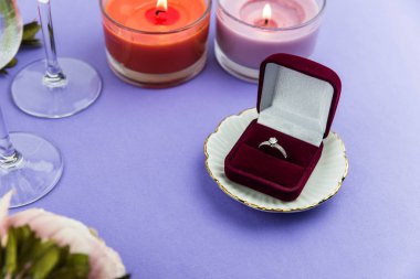 Evlilik teklifi ve Sevgililer Günü için aşk konsepti, içinde evlilik ya da nişan yüzüğü olan parlak bir kutu. Bir bardak şampanya, romantik bir akşam yemeği, şakayık çiçekleri ve modaya uygun mor arka planda mumlar.