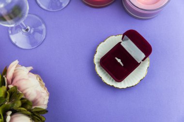 Evlilik teklifi ve Sevgililer Günü için aşk konsepti, içinde evlilik ya da nişan yüzüğü olan parlak bir kutu. Bir bardak şampanya, romantik bir akşam yemeği, şakayık çiçekleri ve modaya uygun mor arka planda mumlar.