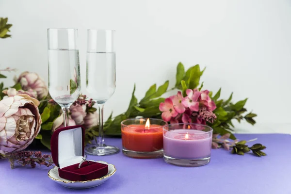 Marriage proposal and love concept for Valentine`s day, box with wedding or engagement ring with brilliant. Glass of champagne, romantic dinner, peony flowers and candles on trendy purple background