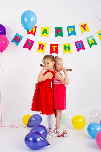 Dos niñas están sosteniendo silbato, de pie junto a la inscripción globos feliz cumpleaños, fondo blanco —  Fotos de Stock