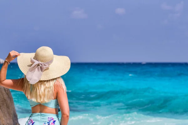 Uma menina em um vestido de verão azul com um chapéu na cabeça, de pé na areia branca e olhando para o oceano — Fotografia de Stock