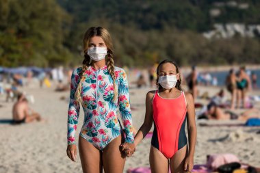 Yüzlerinde koruyucu cerrahi maske takmış bir kadın ve bir çocuk sahilde mayo giymiş duruyor. Çin koronavirüs hastalığı COVID-19 tehlikelidir..