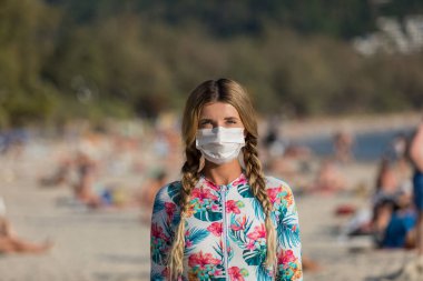 Yüzlerinde koruyucu cerrahi maske takmış bir kadın ve bir çocuk sahilde mayo giymiş duruyor. Çin koronavirüs hastalığı COVID-19 tehlikelidir..