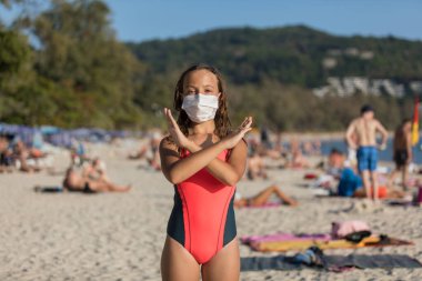 Yüzlerinde koruyucu cerrahi maske takmış bir kadın ve bir çocuk sahilde mayo giymiş duruyor. Çin koronavirüs hastalığı COVID-19 tehlikelidir..