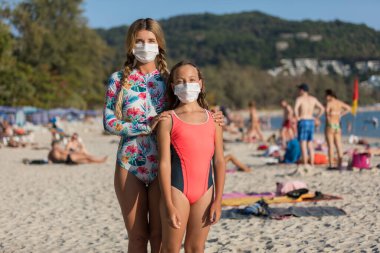 Yüzlerinde koruyucu cerrahi maske takmış bir kadın ve bir çocuk sahilde mayo giymiş duruyor. Çin koronavirüs hastalığı COVID-19 tehlikelidir..