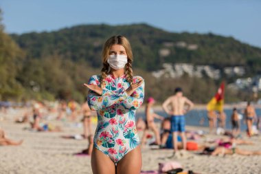 Yüzlerinde koruyucu cerrahi maske takmış bir kadın ve bir çocuk sahilde mayo giymiş duruyor. Çin koronavirüs hastalığı COVID-19 tehlikelidir..