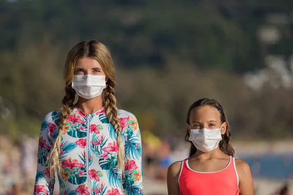 Yüzlerinde koruyucu cerrahi maske takmış bir kadın ve bir çocuk sahilde mayo giymiş duruyor. Çin koronavirüs hastalığı COVID-19 tehlikelidir..